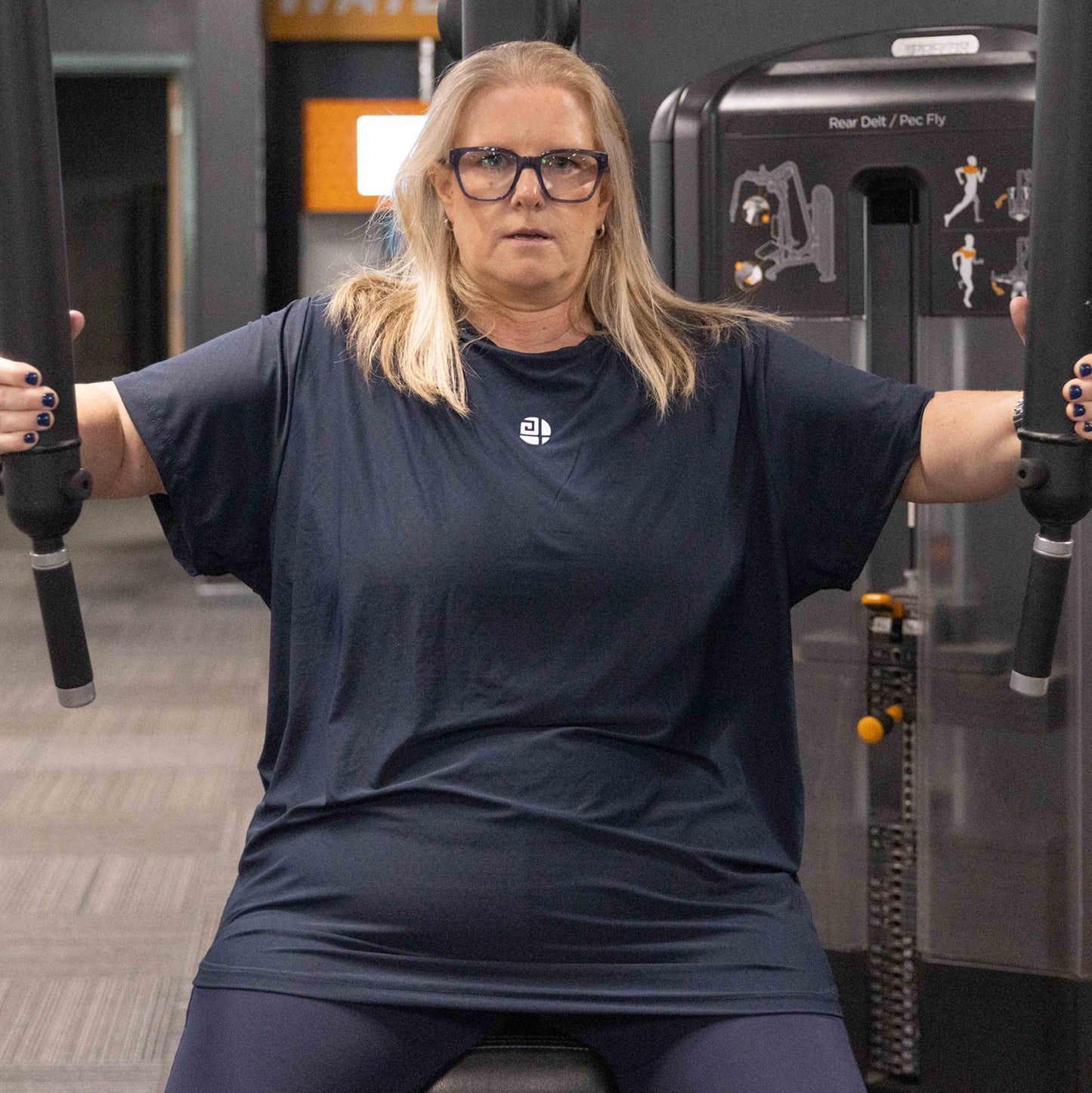 Plus size woman wearing Gym Stuff Plus navy activewear using pec fly gym machine, showing supportive comfortable T-shirt and leggings for real women.