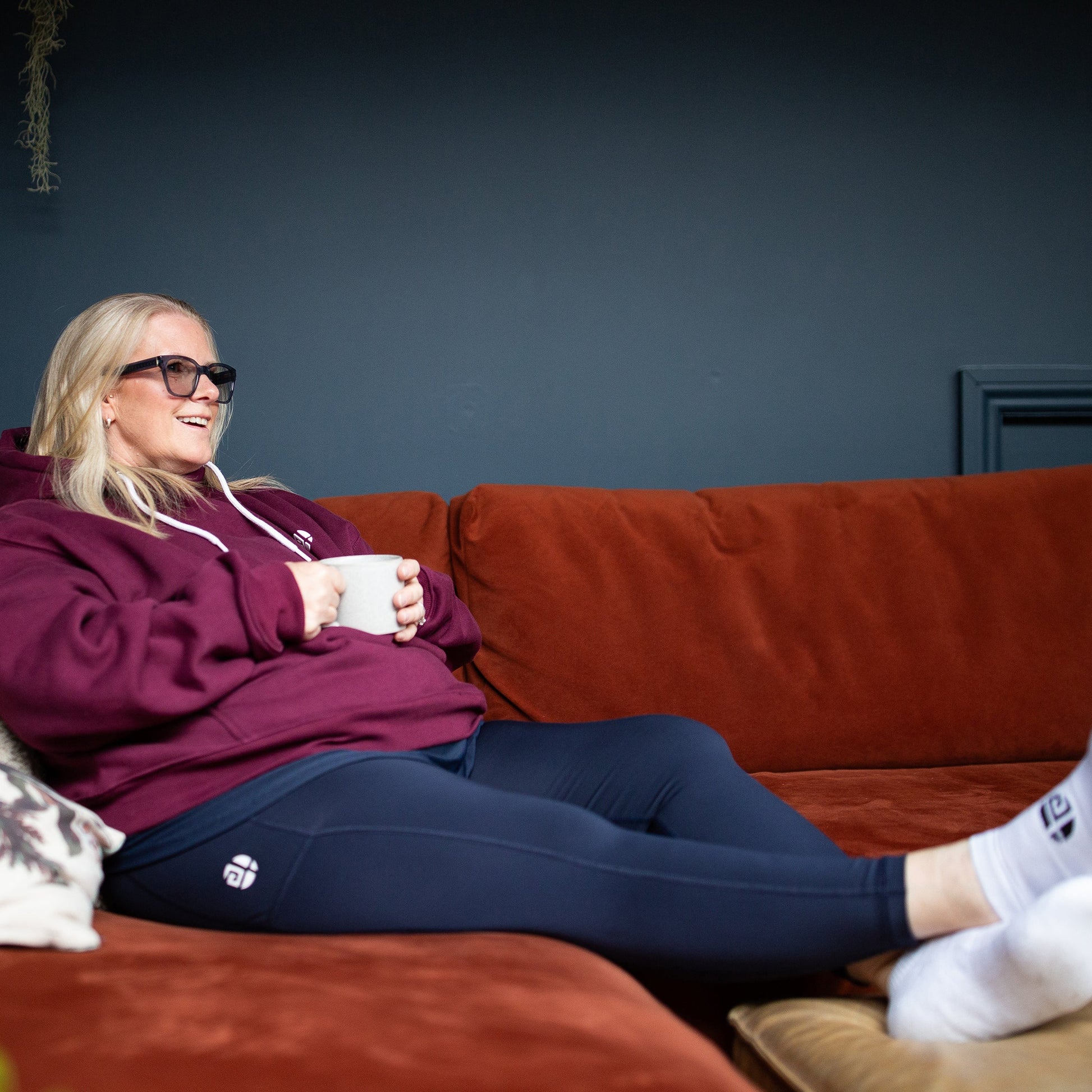 A woman wearing a burgundy hoodie, navy leggings, and white socks relaxes on a rust-colored couch, holding a white mug. The background features a dark blue-gray wall and a blurred green plant in the foreground.